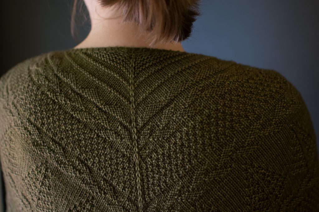 A close up of Ashley's upper back and shoulders. A green shawl with textured patterns resembling mountains, leafy vines, moss, and trees, is draped across her back and shoulders.